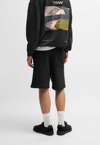 Sweat-shirt noir avec impression graphique au dos, associé à un short noir. Le modèle porte des chaussettes blanches et des sneakers noires. Texture de tissu lisse.