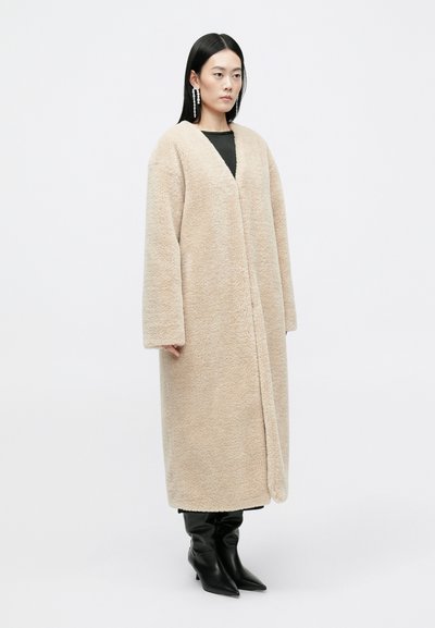 Beige műszőrme kabát puha textúrával, hosszú ujjaival és V-kivágással, fekete felsőre húzva és térdig érő fekete csizmával párosítva.