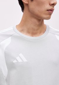 Νεαρό άτομο με ανοιχτό γκρι μπλουζάκι Adidas με λογότυπο στο στήθος, εμφανιζόμενο από τον ώμο μέχρι το πηγούνι σε απλό φόντο.