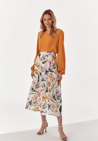 Bluză portocalie cu mânecă lungă și fustă midi cu model floral alb. Fusta prezintă flori vibrante în nuanțe de roz, violet și galben. Tocuri bej cu bretele.