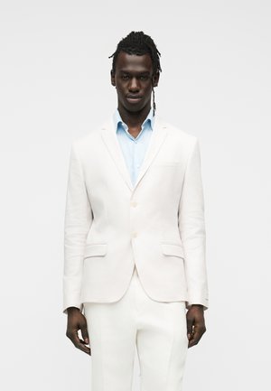 Mann med mørk hud og dreadlocks iført en lyseblå skjorte under en hvit, skreddersydd dressjakke og matchende hvite bukser.