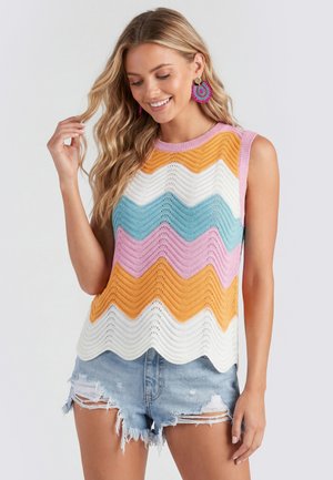 Woman with long blonde hair wearing colorful sleeveless zigzag knit top and distressed denim shorts smiling and touching her hair.