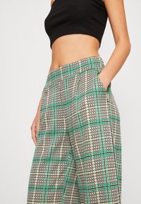 Femme portant un pantalon à carreaux taille haute avec des lignes vertes et noires, associé à un haut noir sans manches cropped, la main dans la poche.