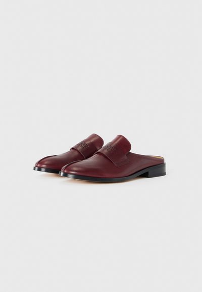 Ett par mörkröda läder slip-on loafers med låga svarta klackar och präglad logotyp på remmen, uppställda mot en vit bakgrund.