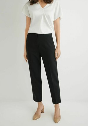 Kobieta ubrana w białą bluzkę z krótkim rękawem i dekoltem w serek, czarne, dopasowane spodnie długości do kostek oraz beżowe buty na słupku, stojąca na tle jednolitego tła.