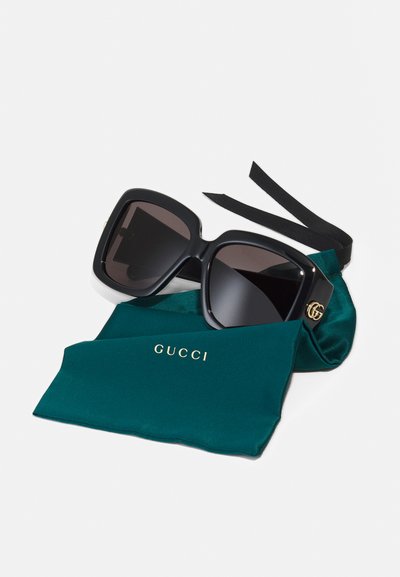 Gucci saulesbrilles ar pārspīlētām melnām taisnstūra rāmjiem un tumšiem stikliem, atpūšoties uz tumši zila mīksta maisiņa ar zelta logotipu un melnām lentēm.