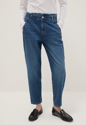 Persona che indossa pantaloni di jeans blu a vita alta con fascia elastica, camicia bianca a maniche lunghe, mani nelle tasche e mocassini neri.