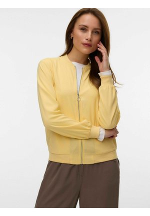 Kobieta o jasnobrązowych włosach, ubrana w żółtą kurtkę na zamek błyskawiczny na białą koszulę i brązowe spodnie, pozując z jedną ręką dotykającą twarzy.