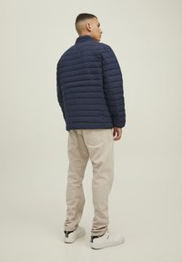 Giacca puffa blu navy con design a cuciture orizzontali, colletto alto, indossata con jeans beige chiari e sneaker bianche. Texture liscia e accenti semplici.