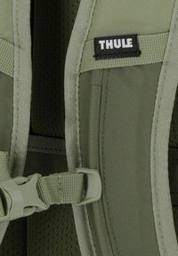 Détail d'un sac à dos en tissu vert avec rembourrage en mesh, bretelles réglables et un logo Thule. Comprend une boucle en plastique et des coutures surpiquées.