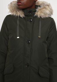Parka verde scuro con cappuccio foderato in pelliccia, dotata di bottoni neri, tasche laterali e cinturini grigi a contrasto. Texture liscia e imbottita.