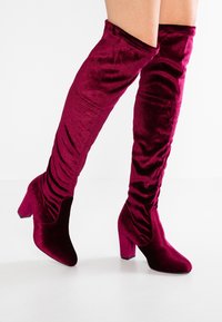 Burgundské sametové nadkolenové boty s elegantním designem, špičatou špičkou a silným podpatkem. Texturovaná tkanina s nařasenými detaily podél hřídele.