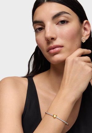 Młoda kobieta o ciemnych włosach, ubrana w czarny bezrękawnik, srebrną bransoletkę z złotym serduszkiem oraz srebrny kolczyk ze złotym detalem.