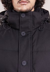 Svart huva-jacka med ett hållbart tyg, stora knappar, justerbar dragsko och strukturerad yta. Designen prioriterar funktionalitet.