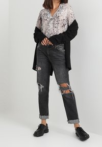 Jean noir déchiré avec ourlets effilochés, associé à un haut boutonné imprimé serpent et une veste noire ample. Des chaussures noires à lacets complètent la tenue.