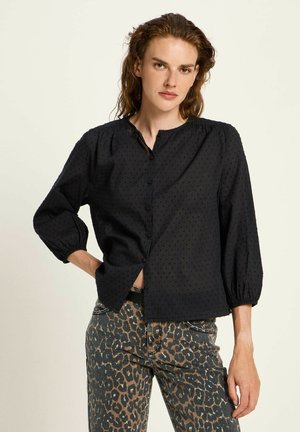 Black blouse with dotted texture, button front, and balloon sleeves. Paired with leopard print jeans, showcasing a relaxed cut and style.