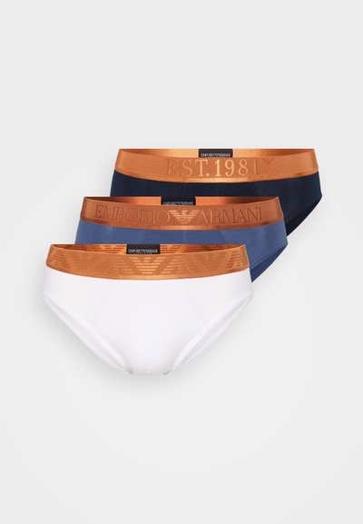 Τρία ζευγάρια ανδρικών εσώρουχων σε ναυτικό μπλε, μπλε και λευκό, με πορτοκαλί ελαστική ζώνη μέσης που έχει ανάγλυφο λογότυπο και λεπτομέρειες της χρονιάς ίδρυσης.