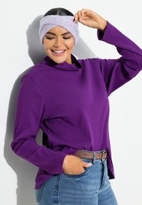 Maglia viola a maniche lunghe con colletto alto, abbinata a jeans azzurri chiari e una cintura marrone. Un morbido cerchietto di colore chiaro completa il look.