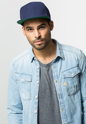 Cappellino navale con ala piatta e parte interna verde, indossato con una camicia in denim azzurro chiaro e una t-shirt grigia. La camicia ha due tasche sul petto.