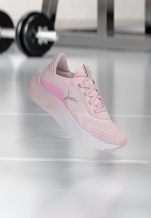 Ένα μόνο ροζ αθλητικό παπούτσι Puma με λευκή σόλα αιωρείται πάνω από λευκό πάτωμα γυμναστηρίου, με θολά βάρη γυμναστηρίου στο παρασκήνιο.