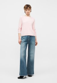 Femme portant un pull à manches longues rose clair, un jean bleu à jambes larges et des bottines noires, debout devant un fond clair uni.