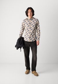 Lys beige skjorte med knapper og blåt geometrisk mønster, sorte jeans og brune slip-on sko. Holder en sort jakke.