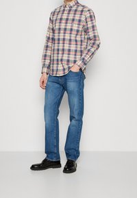 Man staat met één hand in de zak, draagt een meerkleurig geruit overhemd met knopen, blauwe spijkerbroek en zwarte nette schoenen tegen een effen achtergrond.