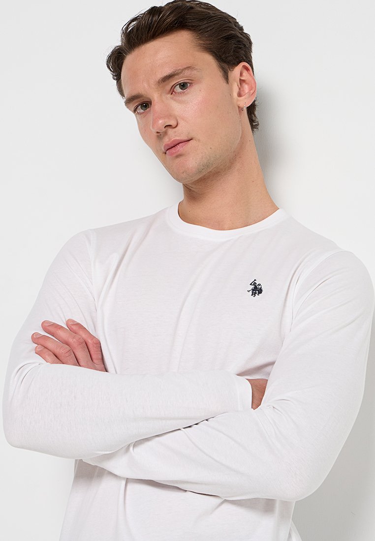 T-shirt bianco a maniche lunghe realizzato in tessuto morbido, con un logo blu navy sul lato sinistro del petto, polsini a coste e colletto rotondo.