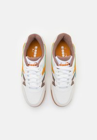 White athletic shoes with perforated detailing, brown and orange accents, teal stripe, and white laces on a brown sole.