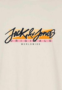 Camiseta beige con un audaz logo en cursiva negro sobre un fondo a rayas colorido en tonos naranja y rosa, que dice "Jack & Jones Originals Worldwide".