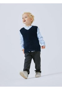 Gilet in maglia blu navy sopra una camicia azzurro chiaro, abbinato a pantaloni grigio scuro e sneakers chiare. Bambino che cammina con un'espressione giocosa.