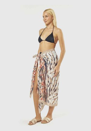 Donna bionda che indossa un top bikini nero e un pareo fantasia legato in vita con sandali beige, in piedi su uno sfondo semplice.