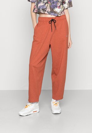 Person trägt lockere, rostorangefarbene Jogginghosen mit schwarzem Kordelzug, ein geblümtes, bauchfreies Oberteil und weiße Nike-Sneaker mit orangen Schnürsenkeln auf grauem Boden.