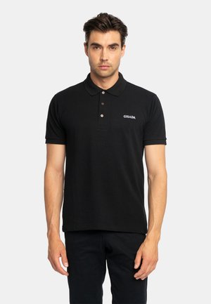 Uomo che indossa una polo nera con tre bottoni e un piccolo logo bianco "GISADA" sul petto, in piedi davanti a uno sfondo chiaro e uniforme.