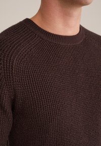 Brūns ribots adīts džemperis ar apaļu kakla izgriezumu un teksturētu audumu. Tuva skatiena izceļ izsmalcināto rakstu un pieguļošo formu.