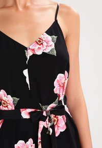 Robe noire à fleurs avec des fleurs roses, des bretelles fines, un décolleté en V et un détail noué à la taille pour une silhouette ajustée.