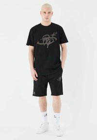 Black short-sleeve t-shirt and shorts set with a large gray graphic design. Worn with white sneakers and socks. Smooth, casual fabric.
