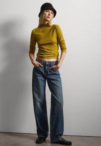 Maglietta gialla a maniche lunghe abbinata a jeans blu larghi. Il modello indossa un cappello a secchiello nero e scarpe nere. Sfondo neutro.