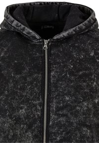 Sweat à capuche noir zippé avec un motif texturé et tacheté, et une fermeture éclair métallique argentée centrée verticalement.