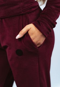 Donkerrode velours joggers met een gestructureerde afwerking, voorzien van zijzakken en een zwart decoratief patch op het onderbeen.