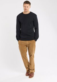 Mann steht mit Händen in den Taschen, trägt einen schwarzen, geknöpften Schulterpullover, khakifarbene Cordhosen und braune Lederschuhe.