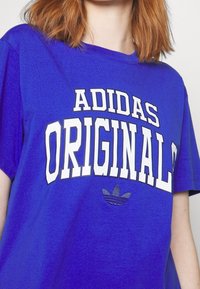Žena nosící jasně modré tričko s bílým textem "ADIDAS ORIGINALS" a černým logem trefoil Adidas na hrudi.