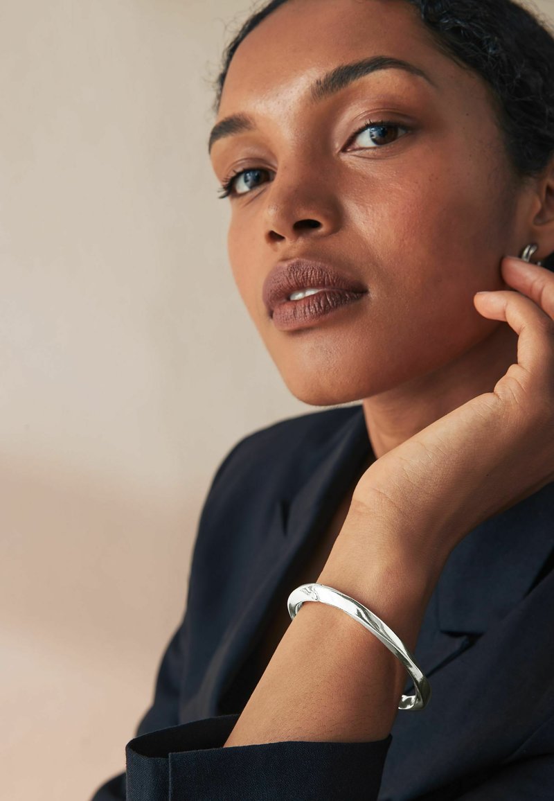 Bracciale d'argento indossato al polso, con una superficie liscia e lucida e un leggero design attorcigliato. Contrasto tra il blazer scuro e le tonalità naturali della pelle.
