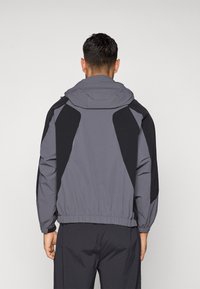 Γκρι μπουφάν με μαύρες λεπτομέρειες, διαθέτει κουκούλα, ελαστικούς σφιγκτήρες στους καρπούς και εφαρμοστή γραμμή. Η πλάτη έχει γωνιώδεις σχεδιαστικές λεπτομέρειες και λεία υφή.