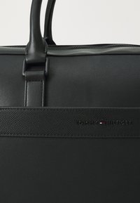 Černá kožená kabelka s hladkou texturou, zaoblenými uchy, zipovým uzávěrem a značkovým akcentem s nápisem "TOMMY HILFIGER" vyraženým tiskem.