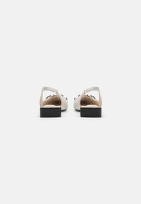 Sandales en cuir blanc avec un talon carré, à dos ouvert et matériel décoratif. Présente une texture lisse et un design minimaliste.