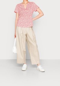 Kortærmet, v-hals top i rød med hvidt blomstermønster. Matcher med beige, vide bukser. Modellen holder en lille hvid håndtaske.