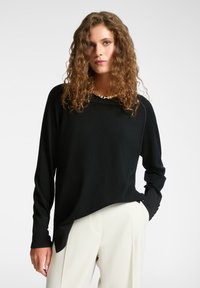Vrouw met krullend haar die een zwarte lange mouwen trui met gouden knopen op de manchetten en off-white broek draagt, poseert met één hand in haar zak.