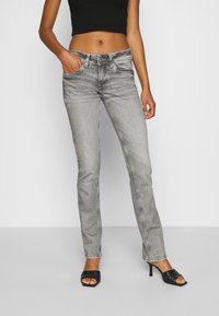 Vaqueros de mezclilla gris ajustados, con diseño de cinco bolsillos, costuras sutiles y cierre de botón. Combinados con tacones abiertos negros.