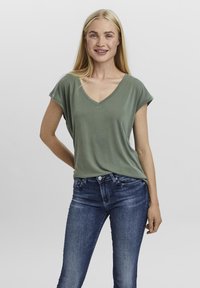 Tricou verde cu decolteu în V, cu mâneci scurte răsucite, asortat cu blugi mulați albaștri. Țesătura pare moale și ușoară, având o textură netedă.